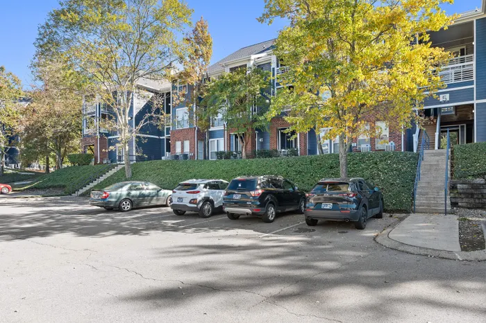 Charming blue apartment complex with modern architecture, ample parking, and lush greenery on a sunny day.