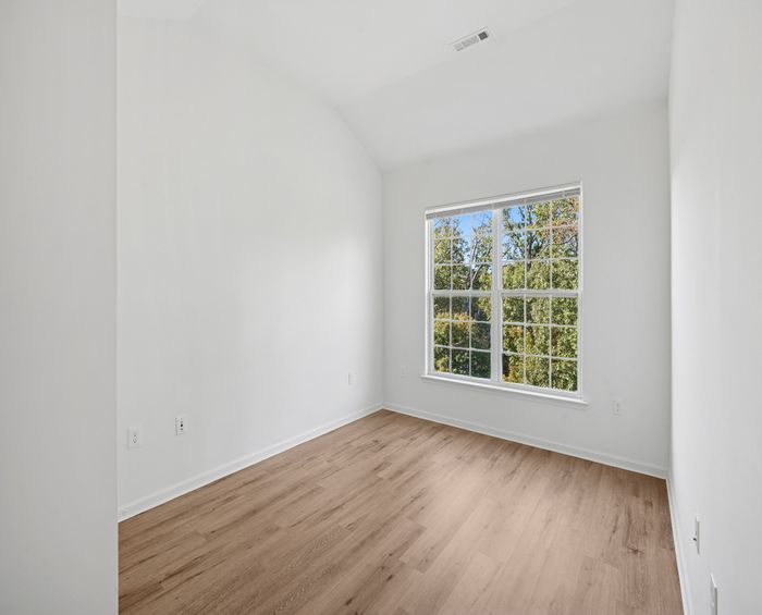 Bright and airy room with large window, elegant wooden flooring, and fresh white walls, offering a serene view of lush trees.