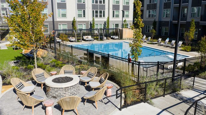 Modern apartment complex courtyard featuring a sparkling pool, cozy fire pit, and elegant lounge seating set amidst lush greenery.
