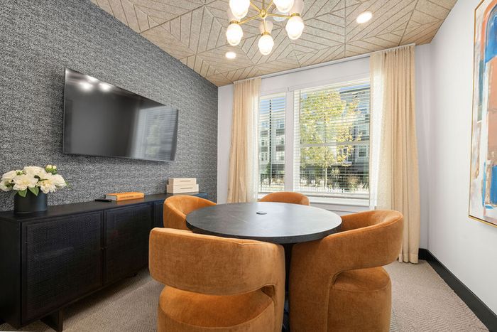 Cozy conference room area with elegant round table, plush velvet chairs, and a large window offering natural light, accented by modern decor and a sleek wall-mounted TV.