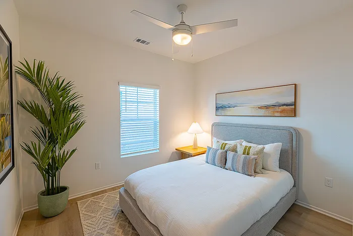 Bright and inviting bedroom featuring a cozy bed with decorative pillows, soft lighting from a side table lamp, stylish ceiling fan, and a vibrant green potted plant, perfect for relaxation.
