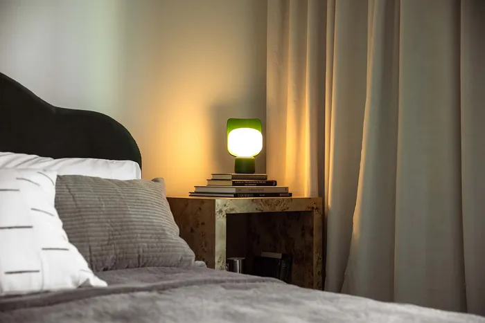 Cozy bedroom corner with a soft bed, elegant side table, and a warm, glowing lamp next to neatly stacked books.