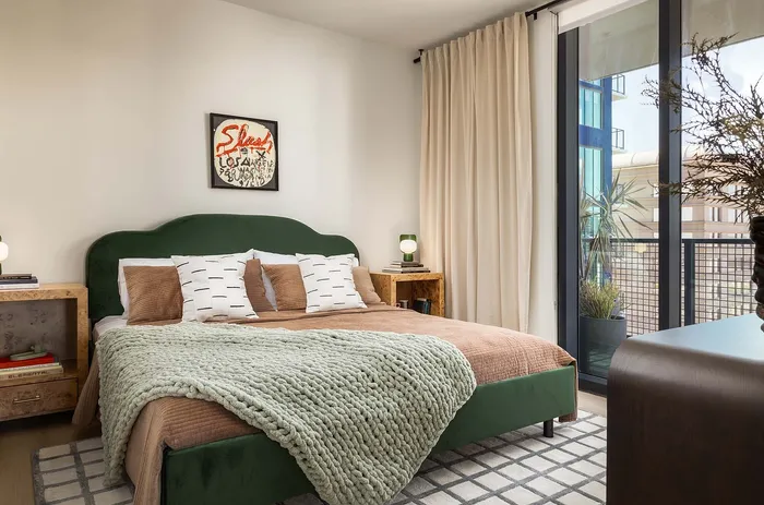 Inviting bedroom with a chic green velvet bed, cozy bedding, modern decor, and abundant natural light from large balcony windows.