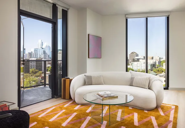 Bright living room with stylish curved sofa, modern glass coffee table, and vibrant geometric rug, featuring expansive city views through large windows.