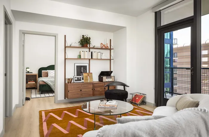 Modern living room with open concept featuring stylish wood shelving, a cozy sofa, and vibrant geometric rug, leading to a chic bedroom with a plush green bed.