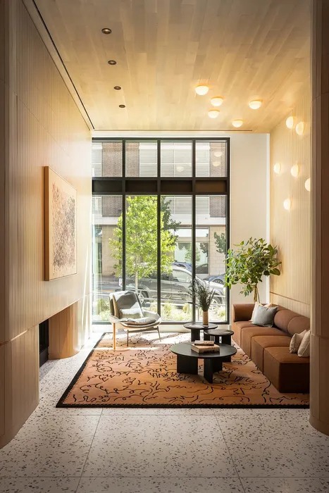 Inviting living room with high ceilings, modern lighting, and floor-to-ceiling windows overlooking lush greenery. Features a stylish brown sofa, cozy armchair, and elegant coffee table arrangement on a patterned rug.