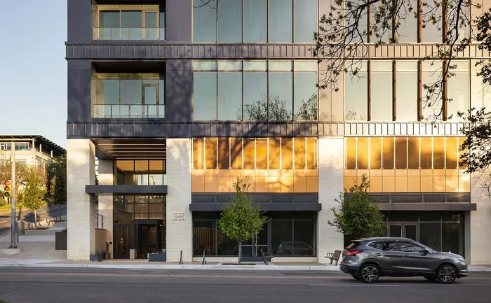 Modern apartment building facade with sleek glass windows reflecting a warm golden hue, complemented by lush greenery and an inviting entrance.