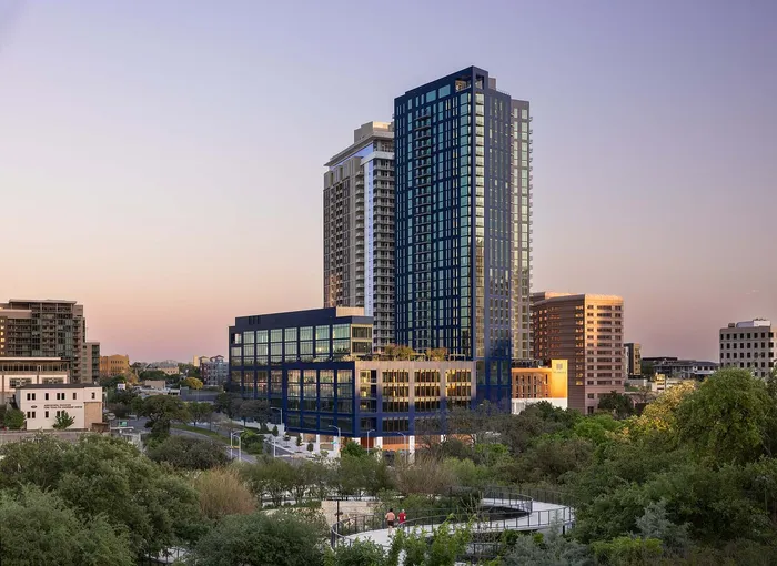 Modern urban high-rise with stunning glass facade, surrounded by lush greenery and a vibrant cityscape at sunset.
