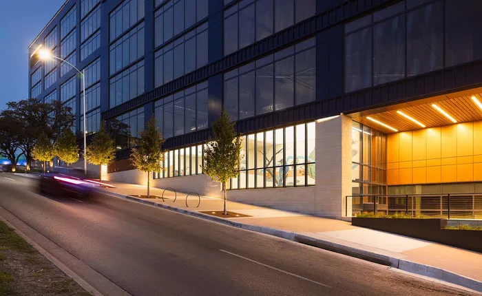 Modern urban apartment building exterior with illuminated windows and sleek architectural design, set on a gently sloping street at dusk.