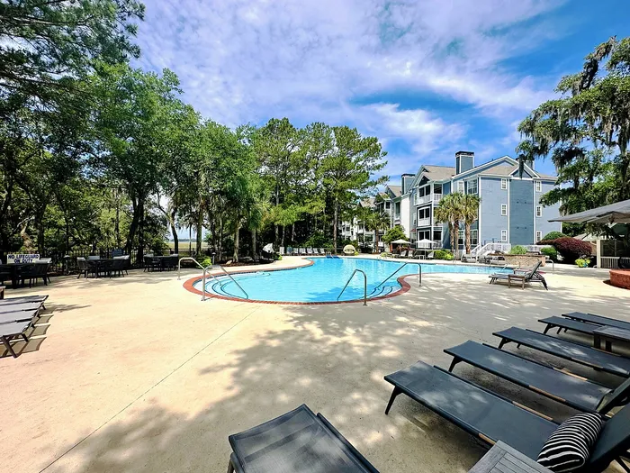 Charming community pool area with relaxing sun loungers surrounded by lush trees and elegant apartment buildings under a brilliant blue sky.