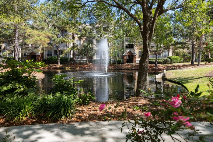 Idyllic apartment complex surrounded by lush greenery, featuring a serene pond with a central fountain and vibrant flowers in the foreground.