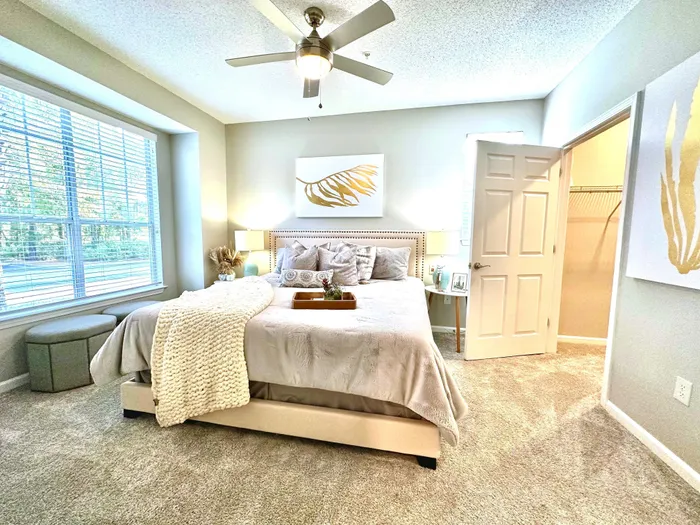 Inviting bedroom with plush carpets, elegant decor, and abundant natural light from large windows, showcasing a cozy bed with stylish bedding and a chic ceiling fan.
