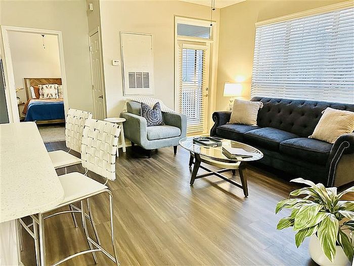 Charming open-concept living area with elegant wood flooring, featuring a cozy blue sofa, stylish armchair, and modern bar seating. Sunlight streams through large windows, enhancing the inviting atmosphere of this apartment.