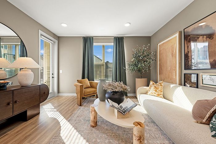 Elegant living room with modern decor, warm beige walls, sunlight streaming through large windows, and stylish furniture featuring a plush sofa and cozy armchair.
