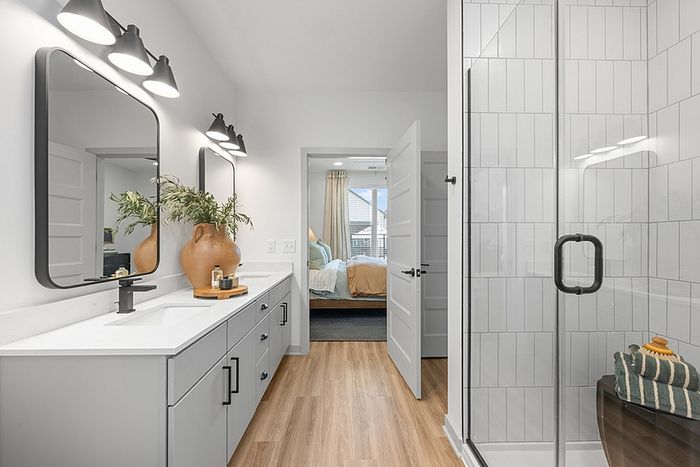 Elegant modern bathroom featuring sleek vanity with double sinks, stylish lighting, and glass-enclosed shower, leading to a cozy bedroom with natural light.