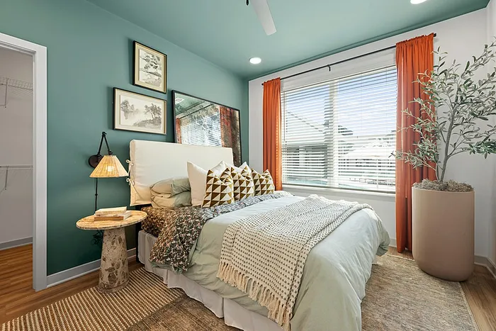 Charming bedroom with teal walls, cozy bed adorned with patterned cushions, and vibrant orange curtains framing a large window for abundant natural light.