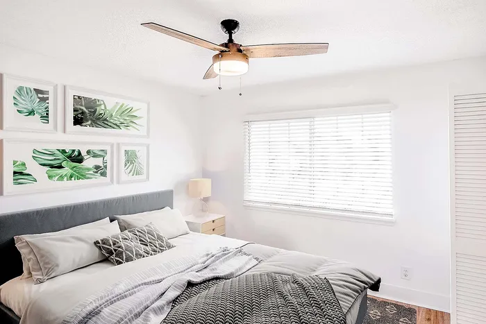 Bright and airy bedroom featuring modern ceiling fan, cozy bed with plush pillows, white blinds, and nature-inspired artwork.