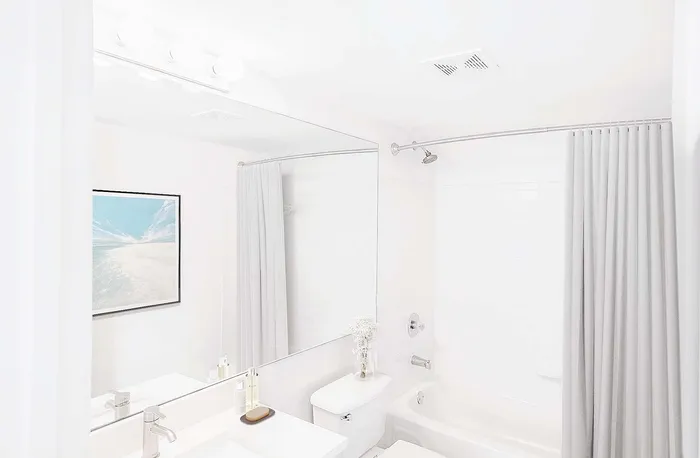 Bright modern bathroom with a large mirror, elegant sink, and a serene framed artwork. Features a white bathtub with a sleek showerhead and soft curtains.