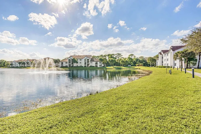 Charming lakeside apartments with red-tiled roofs, surrounded by lush greenery and complemented by a serene fountain under a bright blue sky.