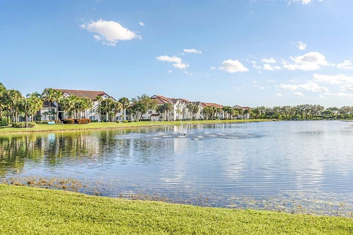 Tranquil lakeside view of elegant apartments surrounded by lush palms and vibrant greenery, under a clear blue sky.