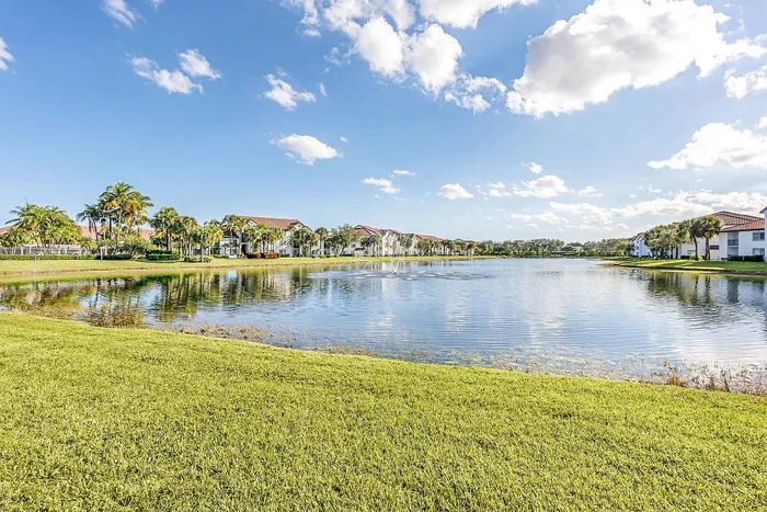 Serene lakeside view with palm trees and charming villas under a bright, blue sky, offering a tranquil living experience.