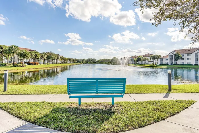 Serene lakefront view with a vibrant bench, lush greenery, and picturesque water fountain, surrounded by charming apartments and palm trees.