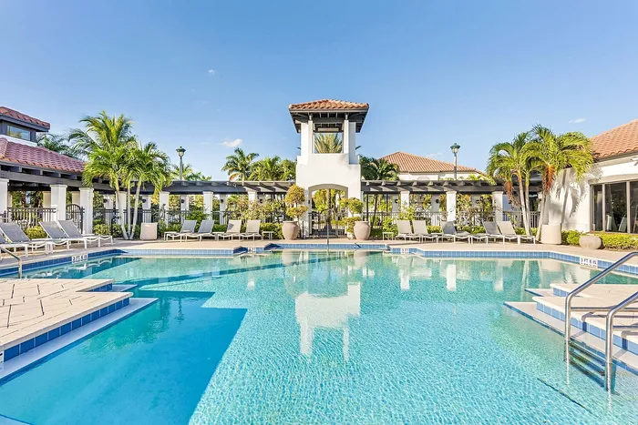 Luxurious swimming pool area with elegant lounge chairs, surrounded by lush palm trees and modern architecture under a clear blue sky.