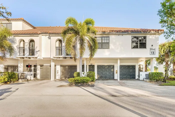Charming two-story townhouse with elegant arches, lush palm trees, and modern garage doors, basking in warm sunlight.