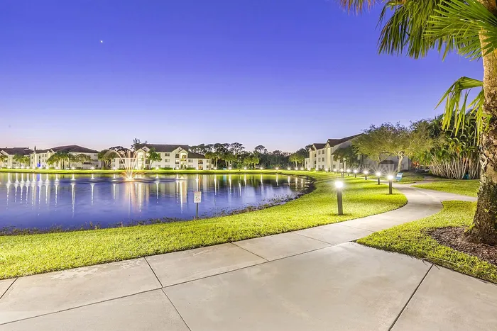 Enchanting twilight view of waterfront apartments surrounded by tranquil lake and lush greenery, highlighted by glowing pathway lights.