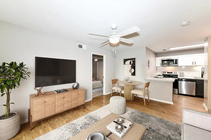 Chic modern living room with a ceiling fan, large flat-screen TV, and light wood flooring adjacent to a stylish kitchen with stainless steel appliances.