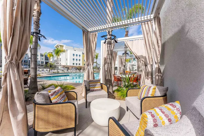 Luxurious outdoor cabana area with stylish wicker seating and vibrant cushions, overlooking a sparkling pool with modern apartment buildings in the background.