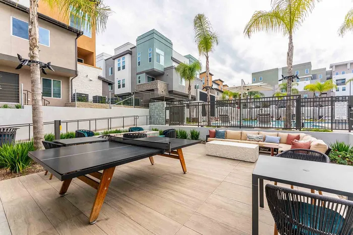Modern outdoor lounge area with sleek poolside seating and a stylish ping pong table, surrounded by contemporary apartment buildings and lush palm trees.