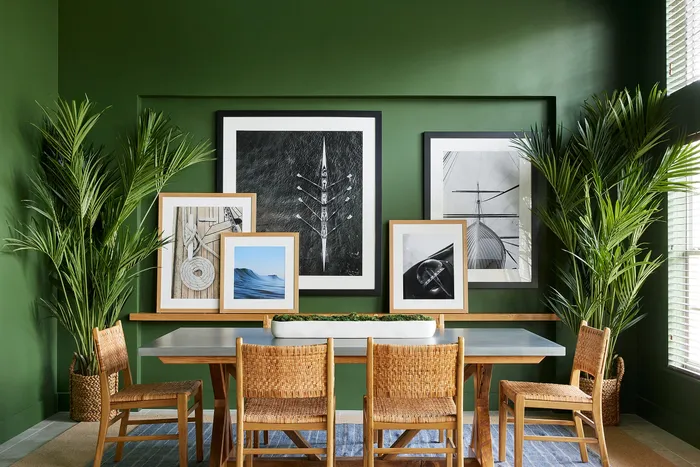 Modern dining area with a sleek table, wicker chairs, and vibrant green walls adorned with framed artworks and lush plants.