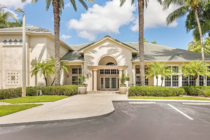 Elegant apartment entrance with Mediterranean-style architecture, lush palm trees, and welcoming pathways under a clear blue sky.