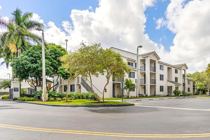 Charming white apartment complex surrounded by lush greenery, featuring elegant balconies and modern architecture under a bright blue sky.