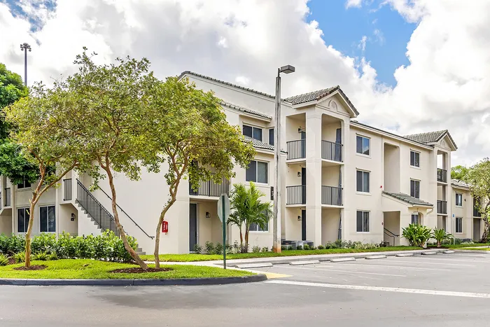 Elegant modern apartment building with lush green landscaping, spacious balconies, and ample parking under a bright blue sky.