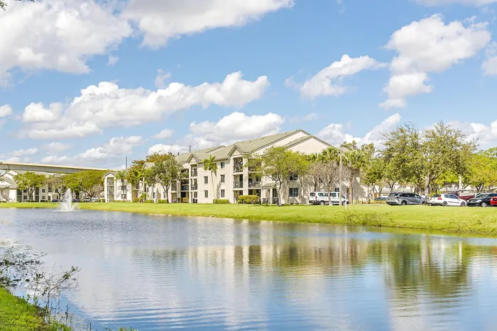 Charming lakeside apartment complex featuring landscaped grounds, serene water views, and inviting architecture under a bright blue sky.