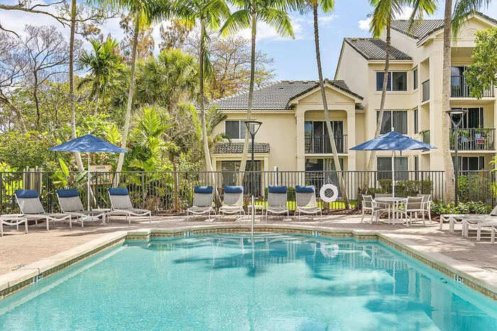 Inviting apartment complex with palm trees and a sparkling outdoor pool, surrounded by sun loungers and cozy seating areas, promising a tropical paradise living experience.