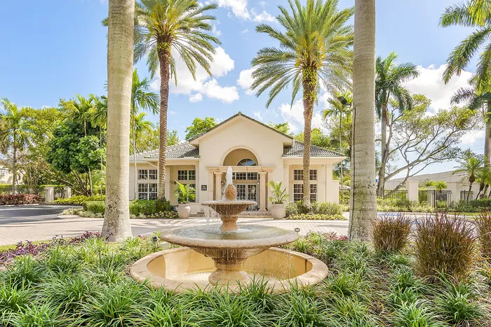 Elegant apartment entrance with lush landscaping and a charming fountain, surrounded by tall palm trees under a bright blue sky.