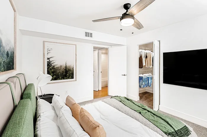 Bright, modern bedroom featuring elegant green accents, a plush bed, stylish ceiling fan, built-in closet, and wall-mounted TV.