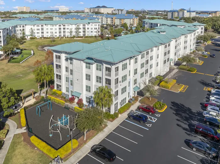Aerial view of a vibrant apartment complex featuring modern teal roofs, manicured landscaping, and a charming playground, surrounded by ample parking space.