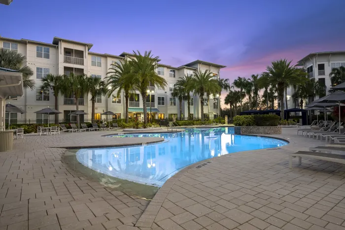Stunning apartment complex featuring a luxurious poolside area with palm trees, sun loungers, and modern architecture against a vibrant sunset sky.