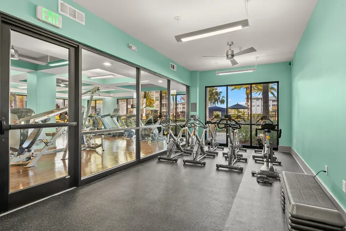 Modern fitness center with sleek exercise equipment, vibrant teal walls, and a view of palm trees through large windows.