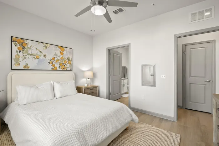 Bright bedroom with a sleek modern design, featuring a cozy bed with white linens, vibrant artwork, and a ceiling fan.