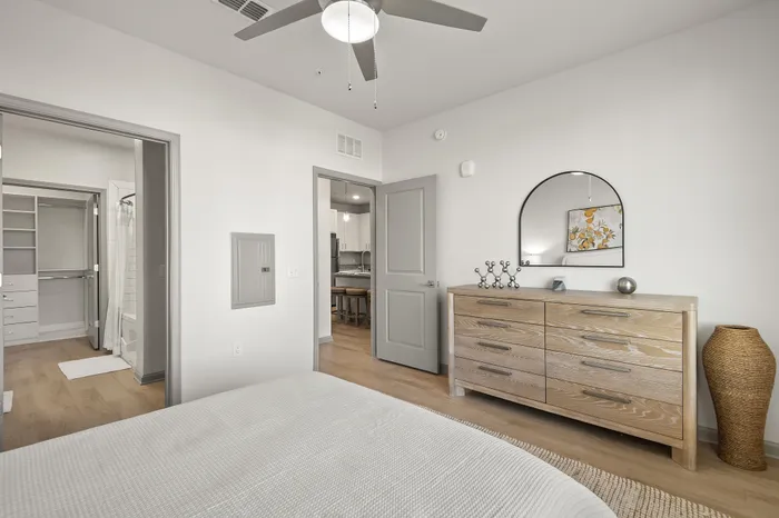 Bright bedroom with stylish decor featuring a modern ceiling fan, chic dresser, and a glimpse into a walk-in closet, complemented by sleek hardwood floors and a neutral color palette.