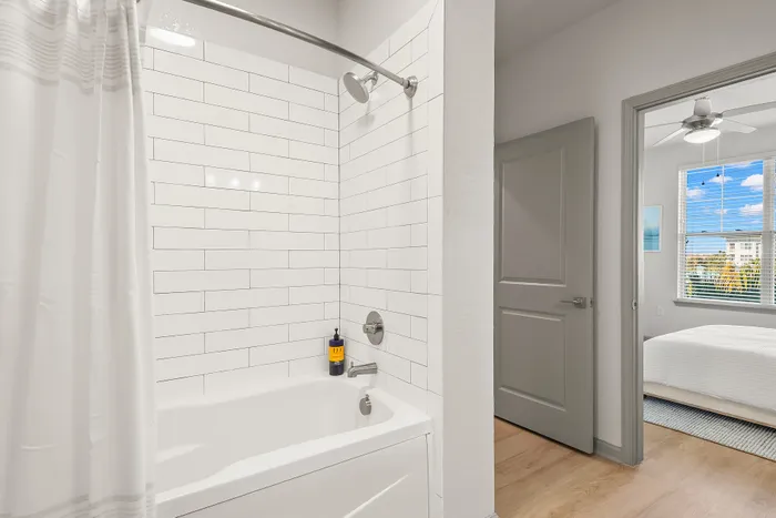 Contemporary bathroom with sleek white tiles, a modern shower, and view into a bright bedroom with large windows.