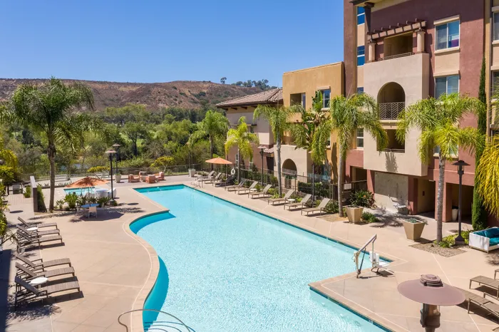Luxurious apartment complex showcasing a sparkling pool surrounded by palm trees and elegant sun loungers, set against a serene natural landscape.