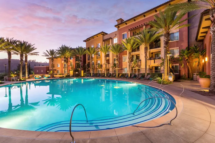 Luxurious apartment complex showcasing a sparkling pool surrounded by lush palm trees and warm ambient lighting at sunset.