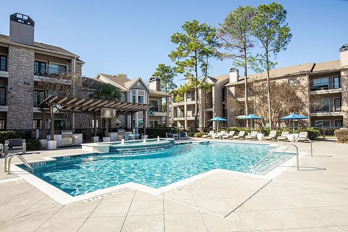 Luxe apartment complex with a sparkling pool, shaded lounge area, and beautifully landscaped surroundings under a sunny sky.