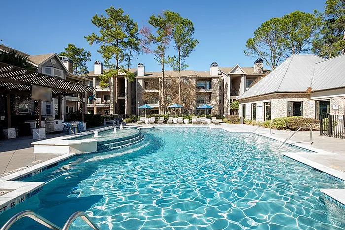 Luxurious apartment complex with a sparkling swimming pool, lounge chairs, and shaded seating against a backdrop of lush greenery.